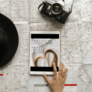 Hand holding a digital wellness guide on hair loss for women, surrounded by travel and lifestyle items on a modern table.