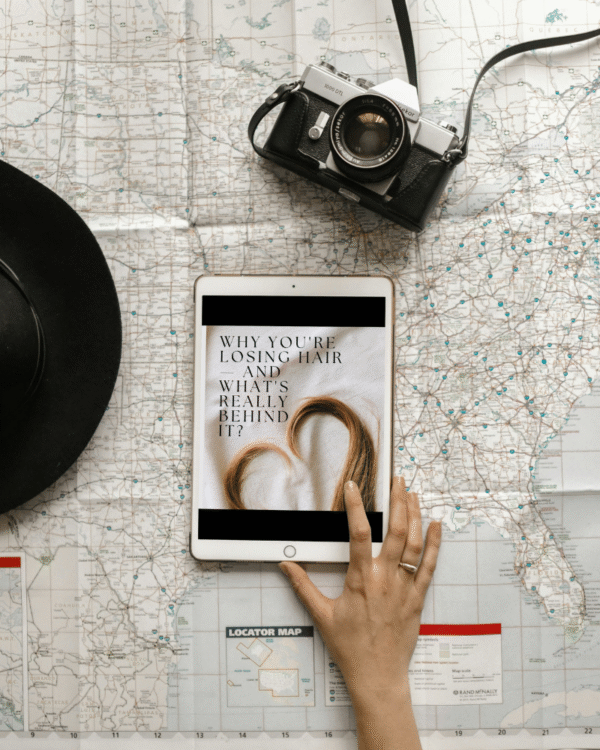 Hand holding a digital wellness guide on hair loss for women, surrounded by travel and lifestyle items on a modern table.